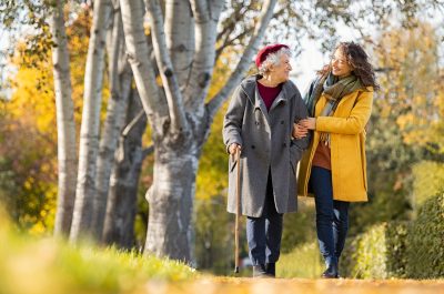 Granddaughter walking with senior woman in park wearing winter clothing. Old grandmother with walking cane walking with lovely caregiver girl in sunny day. Happy woman and smiling grandma walking in autumn park.